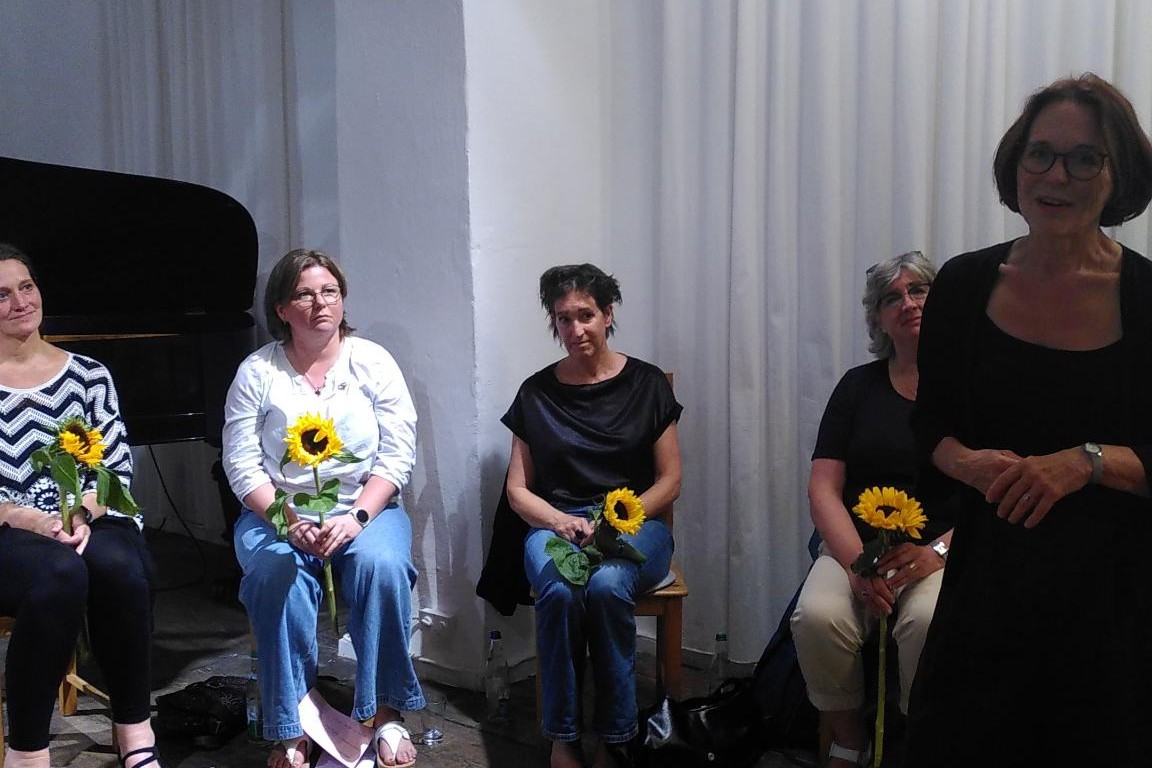 Vier sitzende Frauen mit Sonnenblumen in den Händen hören einer stehenden, vortragenden Frau zu