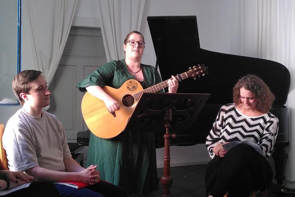 Mittig steht eine Frau mit Gitarre und trägt ein Lied vor, neben ihr sitzt zuhörend links ein Mann, rechts eine Frau